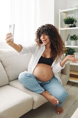 Pregnant woman blogger sits on the couch at home and takes pictures of herself on the phone, selfie and video call, consultation with the doctor online, pregnancy management