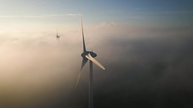 Fog and clouds move between rotating blades of large wind turbines during sunrise
