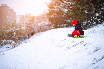 Naklejka na ściany i meble A little boy in a red hat and red pants slides down a snow slide on a round snow board
