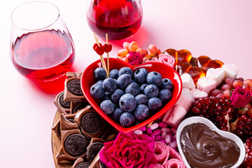 Snack board with berries, chocolate and sweets for Valentine's Day and two glasses of red wine on a pink background
