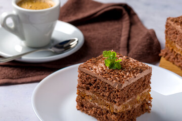 Chocolate cake cut into square pieces with nut cream. Dessert on a white plate with coffee