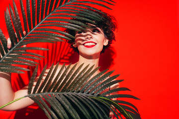 Fashion beauty portrait of young smiling brunette woman with evening stylish  makeup and perfect clean skin. Sexy model with curly short hair posing in studio. With red bright lips. Tropical palm leaf