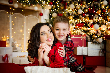 Fototapeta na wymiar smiling happy mother and son wearing red pajamas posing and hugging near christmas tree in decorated living room