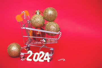 Christmas balls in shopping cart with garland and wooden numbers 2024 on red background.