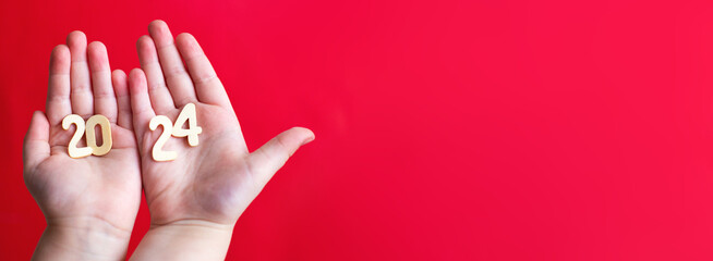 Child hands holding wooden number 2024 on palms on festive red background with copy space