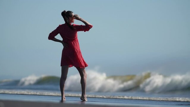 Mature adult woman standing on sandy beach against background of breaking wave of Pacific Ocean. Model in red polka dot baby doll dress, one hand on hip, other raised to sunglasses. Slow motion