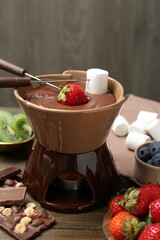Fondue pot with melted chocolate, different fresh berries, kiwi, marshmallows and forks on wooden table