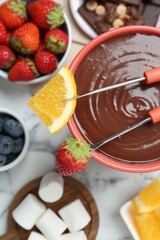 Fondue pot with melted chocolate, marshmallows, fresh orange, different berries and forks on white marble table, flat lay