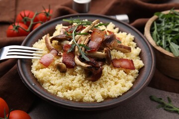 Bowl of tasty couscous with mushrooms, bacon and fork on brown table, closeup
