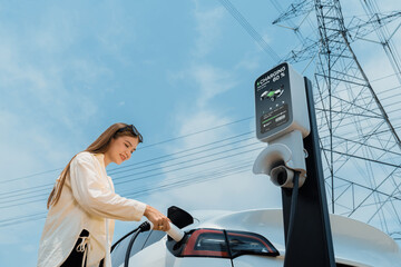 Woman recharge EV electric car battery at charging station connected to electrical power grid tower...