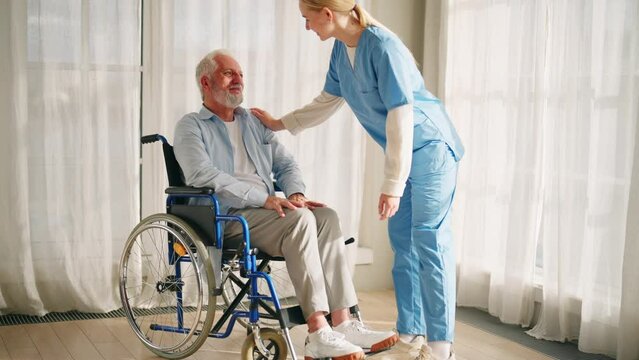 Nurse woman cares old man in wheelchair. Female social welfare worker physical therapist looks after senior male providing medical care at home. Living with legs paralysis, special needs people.