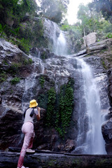 A woman in a hat stands and looks at a waterfall in the forest.