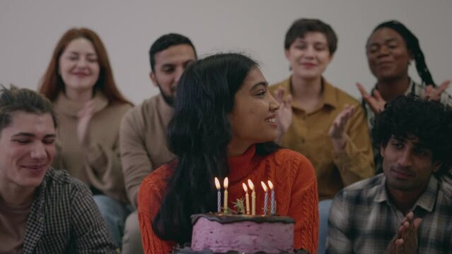 Portrait of attractive indian girl blowing out fire candles on birthday cake. Friends smiling, laughing, rejoicing, congratulating and clapping birthday girl. International friendship, female friend