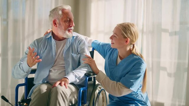 Nurse talking senior man in wheelchair. Female social welfare physical therapist looks after senior male, listening, providing medical care at home. Living with legs paralysis, special needs people.