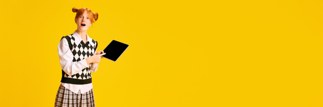 Banner. Adorable, pleasantly shocked young girl, student pointing on tablet with blank screen against yellow background with negative space for text.
