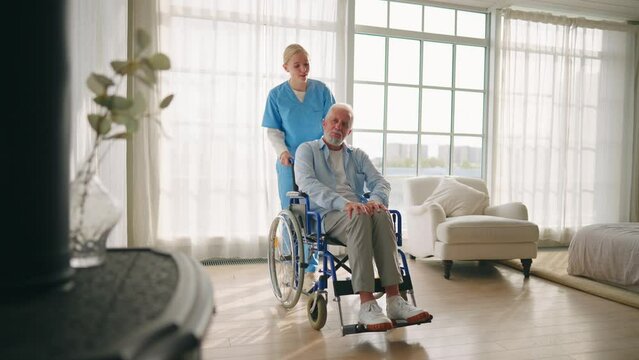 Nurse woman cares old man in wheelchair. Social medical worker pushing wheelchair with paralyzed senior male helping supporting him at home. Living with spine legs paralysis, special needs people.