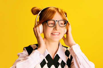 Close up portrait of attractive young girl, student in glasses widely smiling while listening favorite music in headphones against yellow background.