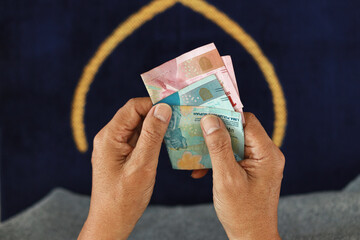 Muslim's hand holding cash money in Indonesia rupiah on prayer mat. Islamic zakat or donation...