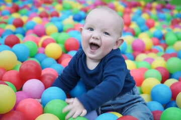 Fototapeta na wymiar A satisfied baby sits among bright multi-colored balls and smiles joyfully. Cute baby in the children's room. Portrait.