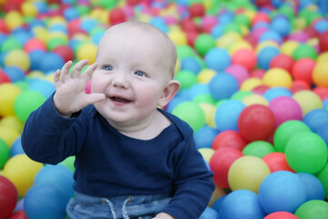 Fototapeta na wymiar A cute eared baby among the colorful balls smiles and waves his hand. Cute baby in the children's room. Close-up portrait