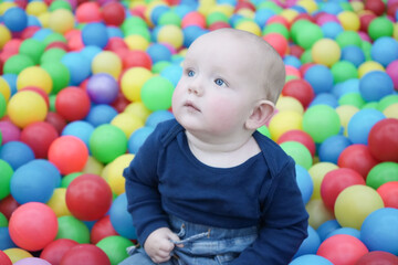 Fototapeta na wymiar A cute eared baby sits among bright multi-colored balls and looks mysteriously to the side. Cute baby in the children's room. Close-up portrait