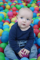 Fototapeta na wymiar A cute eared baby sits among bright multi-colored balls and mysteriously looks up to the side. Cute baby in the children's room. Portrait. Vertical photo.