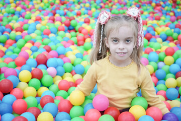 Fototapeta na wymiar A beautiful blond girl with two ponytails sits among colorful bright balls. Close-up portrait. Cute happy girl in the playroom.