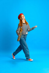 Happy cheerful woman, dressed retro fashion outfit in red beret walking with baguette against blue...