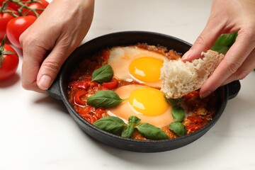 Woman dipping piece of bread into delicious Shakshuka at white table, closeup