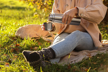 Picnic time. Woman pouring tea from thermos into cup lid on green grass outdoors, closeup