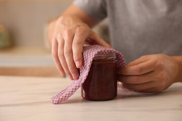 Man packing jar of jam into beeswax food wrap at light marble table indoors, closeup - obrazy, fototapety, plakaty