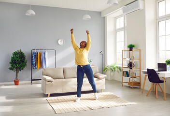 Happy cheerful young African American woman in casual clothes enjoying free time at home, standing in living room with sofa, raising hands up, stretching, smiling and charging with positive energy - obrazy, fototapety, plakaty