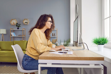 Beautiful young woman accountant is working from home, sitting at her desk, and working with financial spreadsheets on a modern desktop computer. Side view. Remote work concept