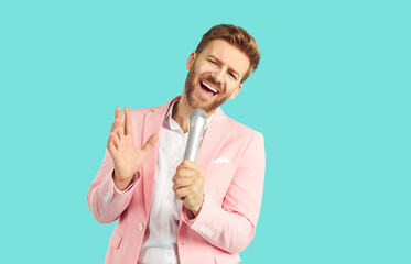 Handsome emotional young man singing over microphone and gesturing. Attractive bearded man wearing elegant pastel pink jacket suit performing isolated on light blue color studio background