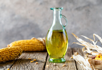 Corn oil in an oil jug with maize cobs over rustic wooden table and grey background