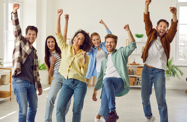 Fototapeta premium Funny happy overjoyed group of friends celebrating success holding hands up and looking at the camera at home. Group of smiling people students in casual clothes standing together enjoying party.