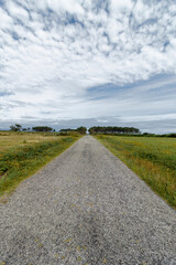 A gravel path leads through a green field towards the horizon, under a cloudy blue sky with trees in the background