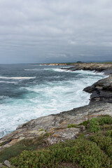 A turbulent ocean crashes against a rocky coastline under a cloudy gray sky, with a small island visible in the distance