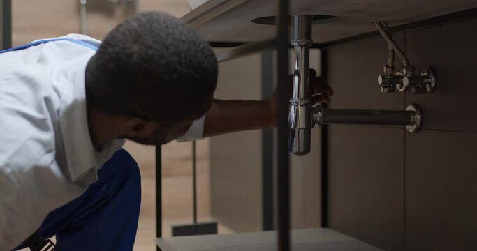 Young plumber at work under kitchen sink, repairing and installing. at home indoors. black man in uniform work in kitchen under sink. He repair water leaking. unscrew the screws under sink