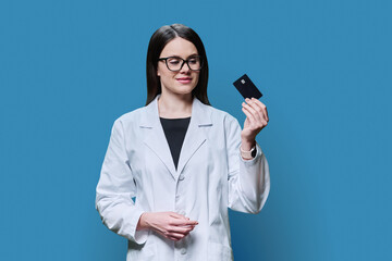 Young woman medical specialist, doctor holding bank credit card on blue background