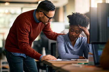 Caring businessman consoling his distraught black female colleague while she's working on desktop PC in office.