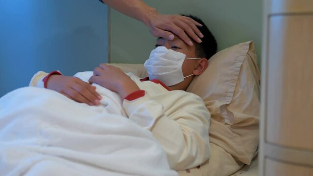 Asian boy wearing a mask was sick and coughing covered in blanket is lying on couch while his father is taking his temperature