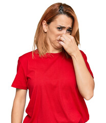 Hispanic young woman wearing casual red t shirt smelling something stinky and disgusting,...
