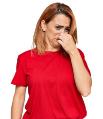 Hispanic young woman wearing casual red t shirt smelling something stinky and disgusting, intolerable smell, holding breath with fingers on nose. bad smell