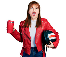 Redhead young woman holding motorcycle helmet and take away coffee angry and mad screaming frustrated and furious, shouting with anger looking up.