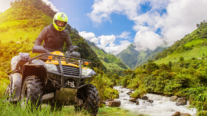 Motorcyclist on quad bike. Man rides ATV off-road. Extreme racing in mountains. ATV racer under summer sky. Quad cyclist in yellow helmet. Driver of quad bike crosses river. ATV near stormy river. - obrazy, fototapety, plakaty