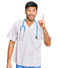 Young handsome man wearing doctor uniform and stethoscope pointing finger up with successful idea. exited and happy. number one.