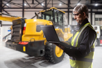 Construction company employee. Man builder with laptop. Construction company engineer. Guy in hangar with bulldozer. Concept development software for construction equipment. Man in reflective vest
