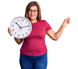 Middle age latin woman holding big clock smiling happy pointing with hand and finger to the side