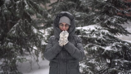A young woman stands against a background of fir trees under heavy snowfall and rubs her hands...